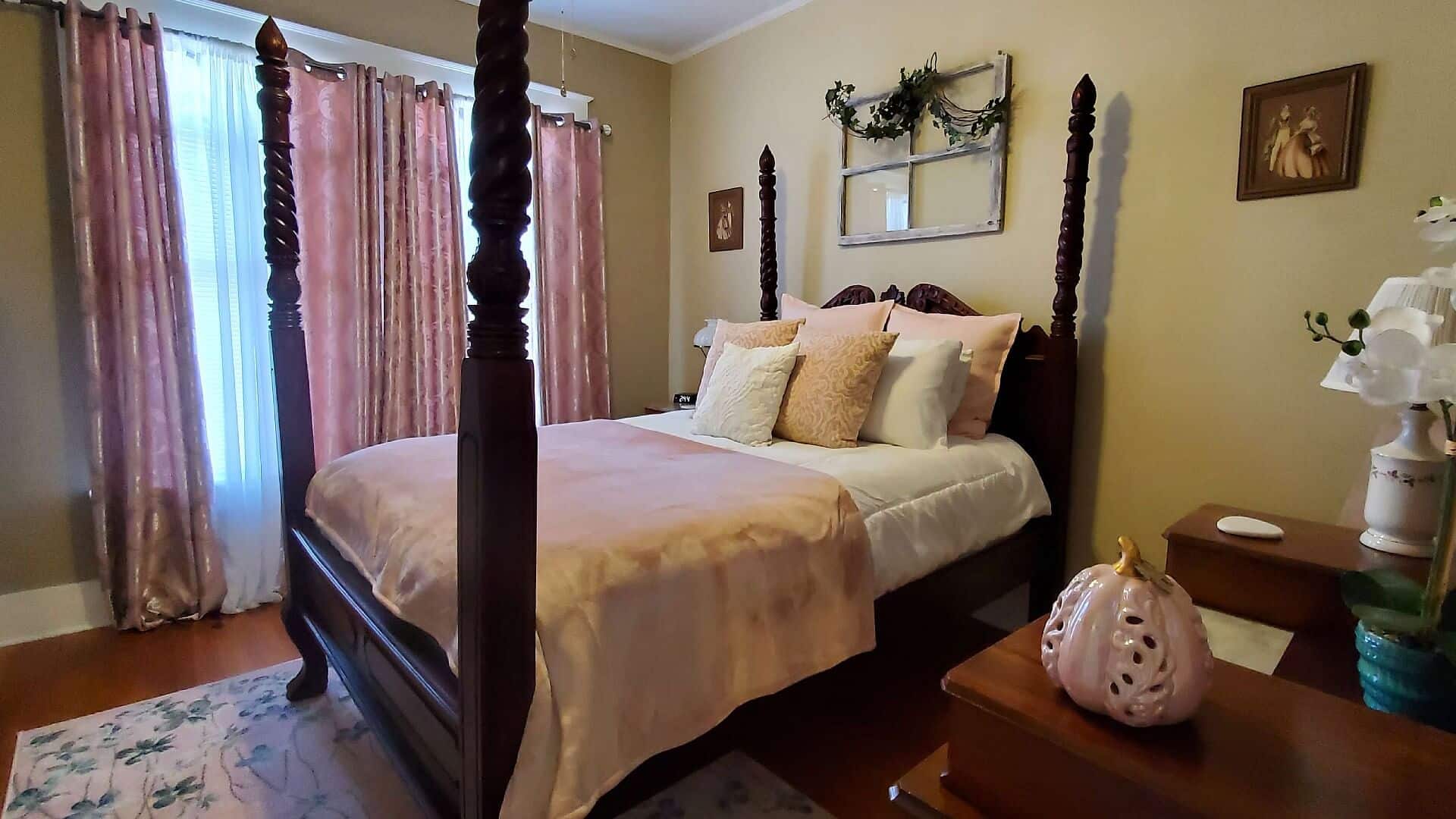 Elegant bedroom with four poster mahogany bed in pink and white linens with an antique dresser and large windows with curtains